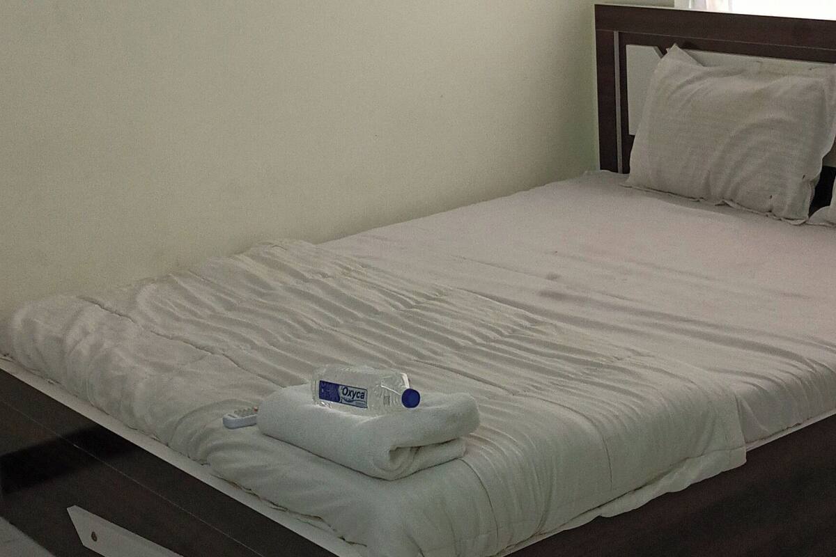 A bedroom with a bed, an air conditioner, a window with curtains, and a bedside table with a bottle.