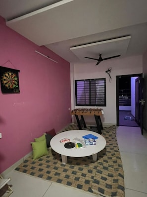 A room with a ceiling fan, a dartboard, a table with various items, and a window with blinds. - Renee Mysaa (Indore)