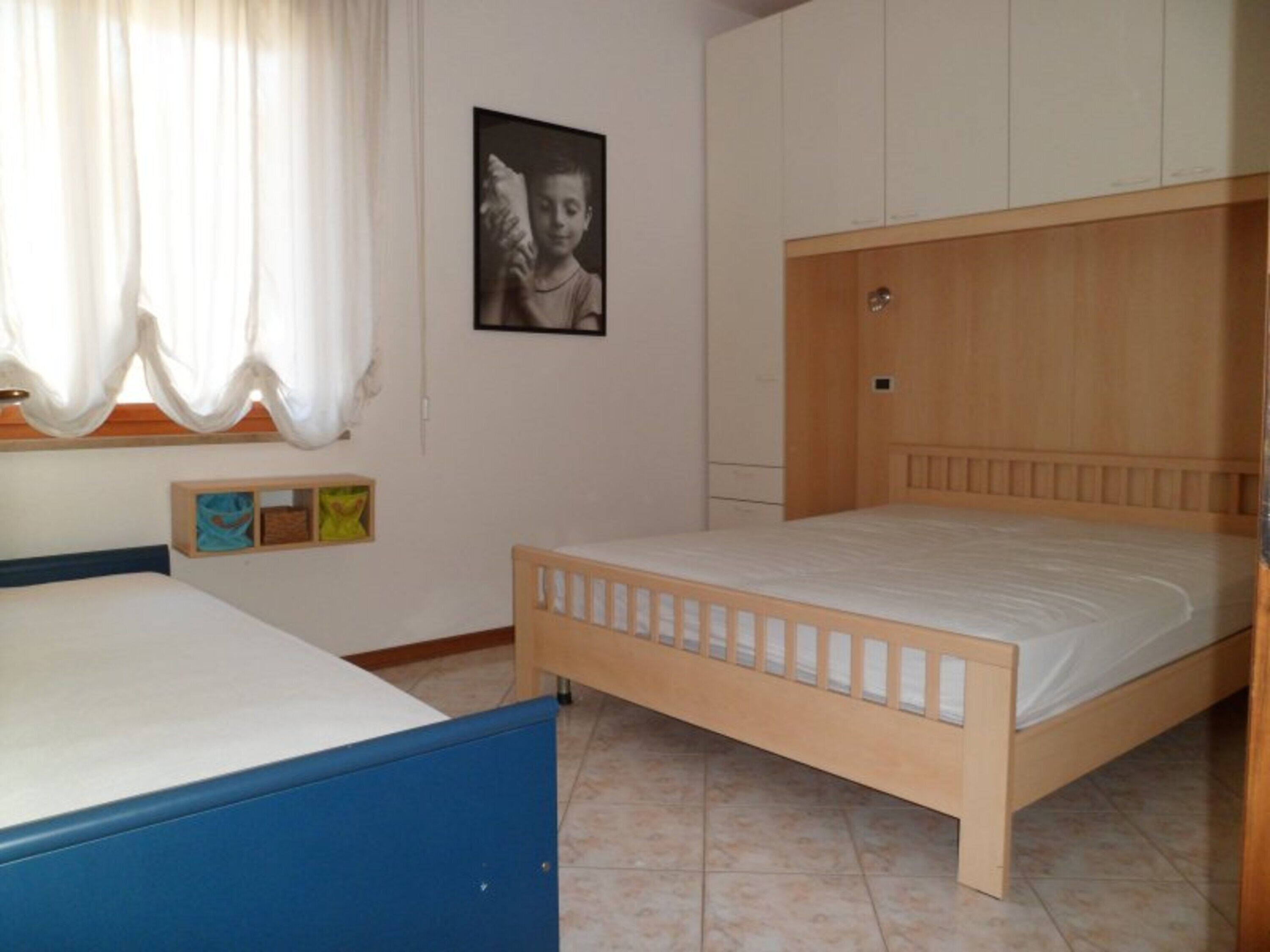 A bedroom with a single bed, a wooden headboard, a window with curtains, and a wall-mounted picture.