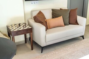 A living room with a white sofa, brown cushions, a chessboard, and a small wooden side table. - Modern Studio l Huge Balcony l New Bldg (Athens)