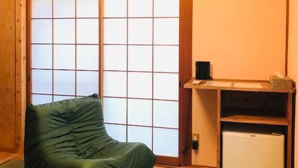 Een kamer met tatami vloerbedekking, een futon, een groene stoel, een kleine tafel en een witte koelkast.