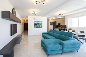 A modern living room with a teal sofa, a flat-screen TV, and a dining area with a table and chairs. - Enjoy a bright 3-bedroom, 2-bathroom apartment on the 4th floor with lift access in St Paul’s Bay. Featuring a spacious living area, modern kitchen, balcony/terrace, sea views, and air conditioning, it is ideal for families and groups. (St. Paul's Bay)