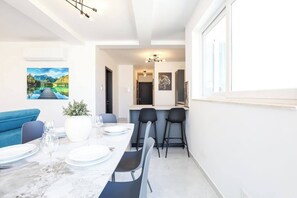 A dining area with a table set for four, a blue sofa, and a painting on the wall. - Enjoy a bright 3-bedroom, 2-bathroom apartment on the 4th floor with lift access in St Paul’s Bay. Featuring a spacious living area, modern kitchen, balcony/terrace, sea views, and air conditioning, it is ideal for families and groups. (St. Paul's Bay)