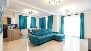 Een moderne woonkamer met een blauwgroene modulaire bank, een keuken met een marmeren aanrecht en grote ramen met blauwgroene gordijnen.