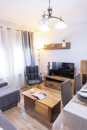 A modern living room with a sofa, a wooden coffee table, a TV, and a dining area with a table set for two. - Vrbo Property (Torrijos)