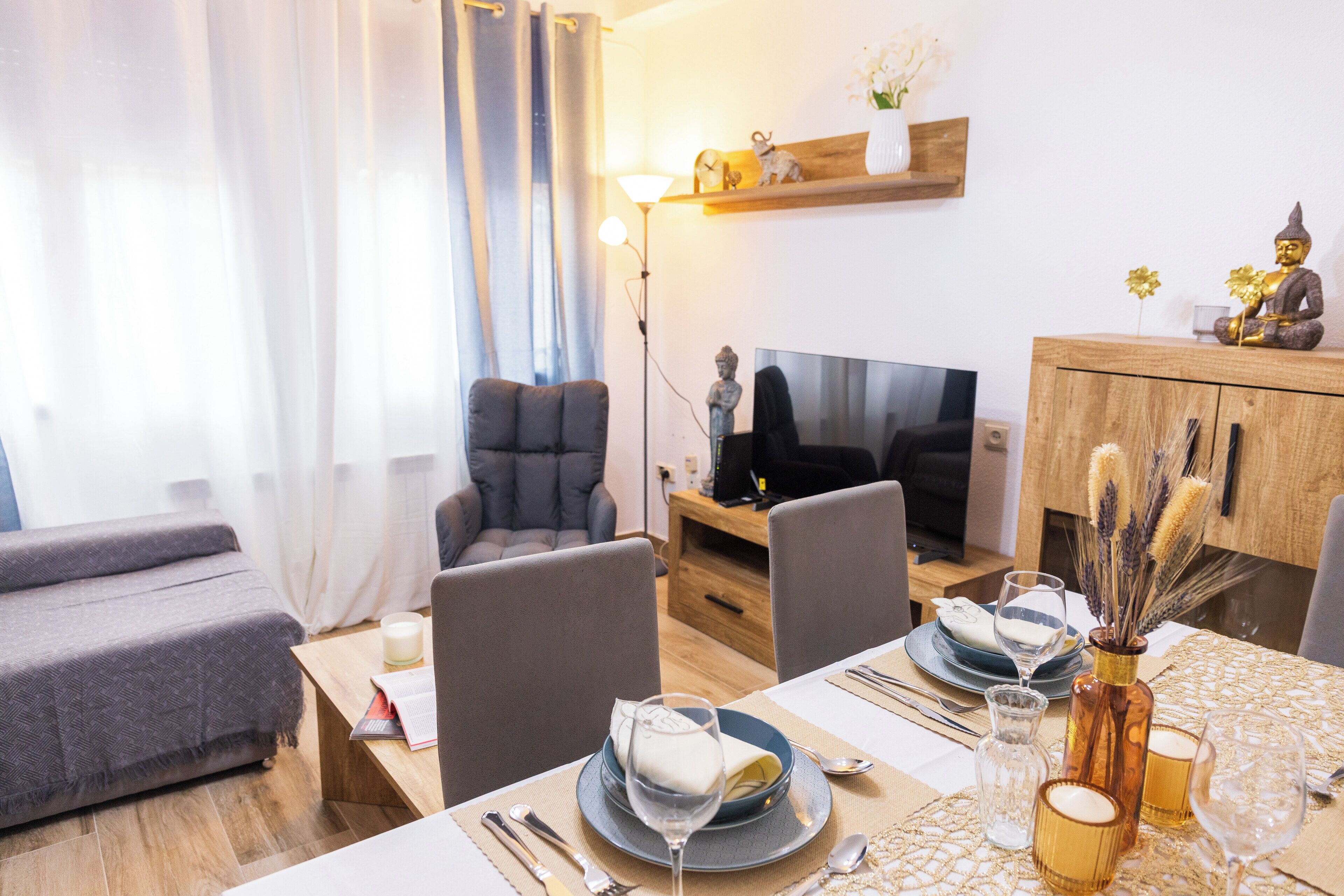 Een moderne woonkamer met een bank, stoelen, een televisie en een eethoek met een tafel gedekt voor twee personen.