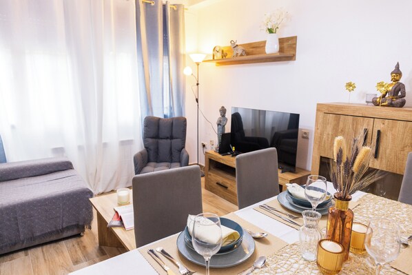 A modern living room with a sofa, chairs, a TV, and a dining area with a table set for two.