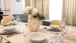 Een eettafel gedekt met witte borden, kommen en wijnglazen, een vaas met witte bloemen, en een bank op de achtergrond.