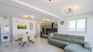 Een moderne woonkamer met een bank, eettafel, stoelen, een wasmachine en een keuken op de achtergrond.