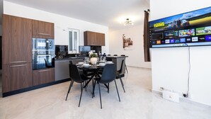 Une cuisine moderne dotée d’appareils électroménagers intégrés, un coin repas avec une table dressée pour quatre personnes, et un téléviseur fixé au mur qui affiche l’interface d’un service de diffusion en continu.