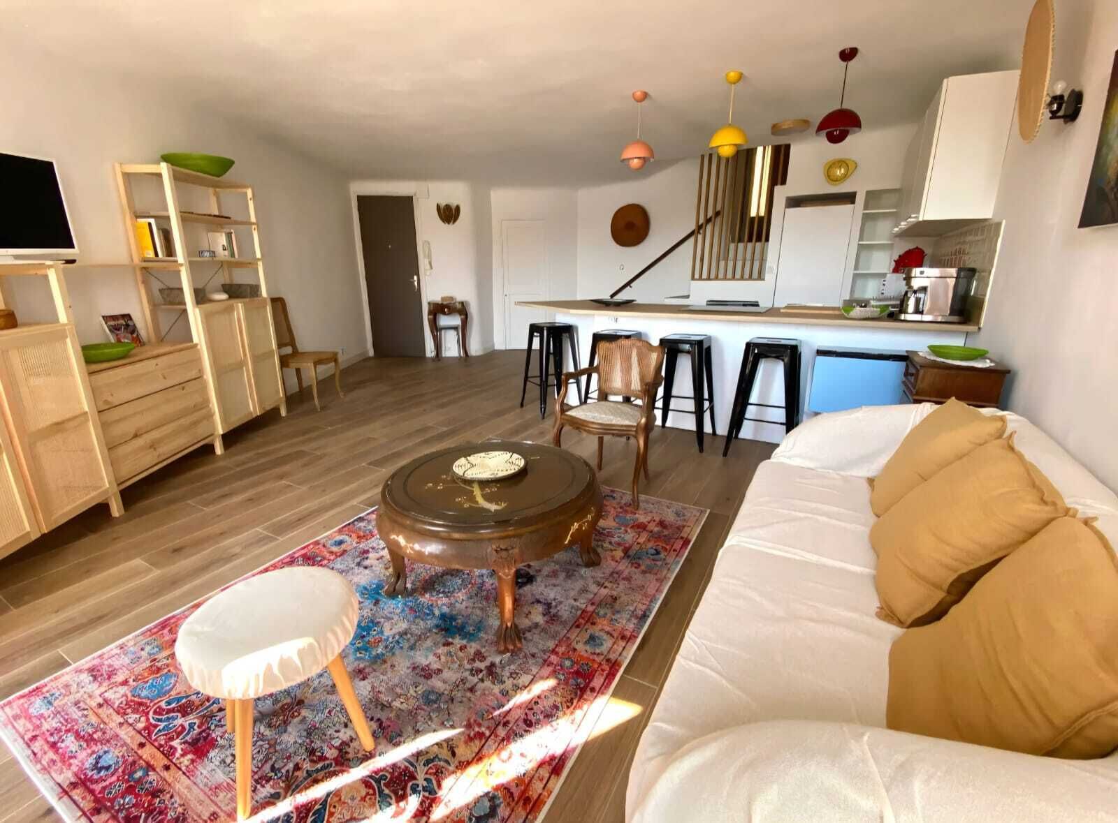 Una sala de estar con un sofá, una mesa de centro y una alfombra. Cuenta con una zona de cocina con mostrador y taburetes.