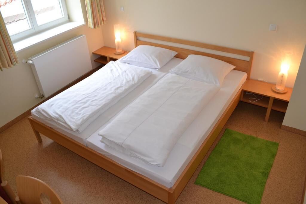 Una cama doble con ropa blanca, dos mesitas de noche con lámparas, un radiador y una alfombra verde.