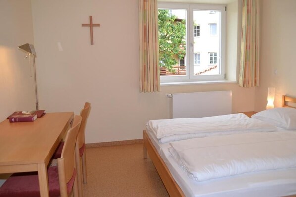 A room with a bed, a desk with a chair, a cross on the wall, and a window with curtains.