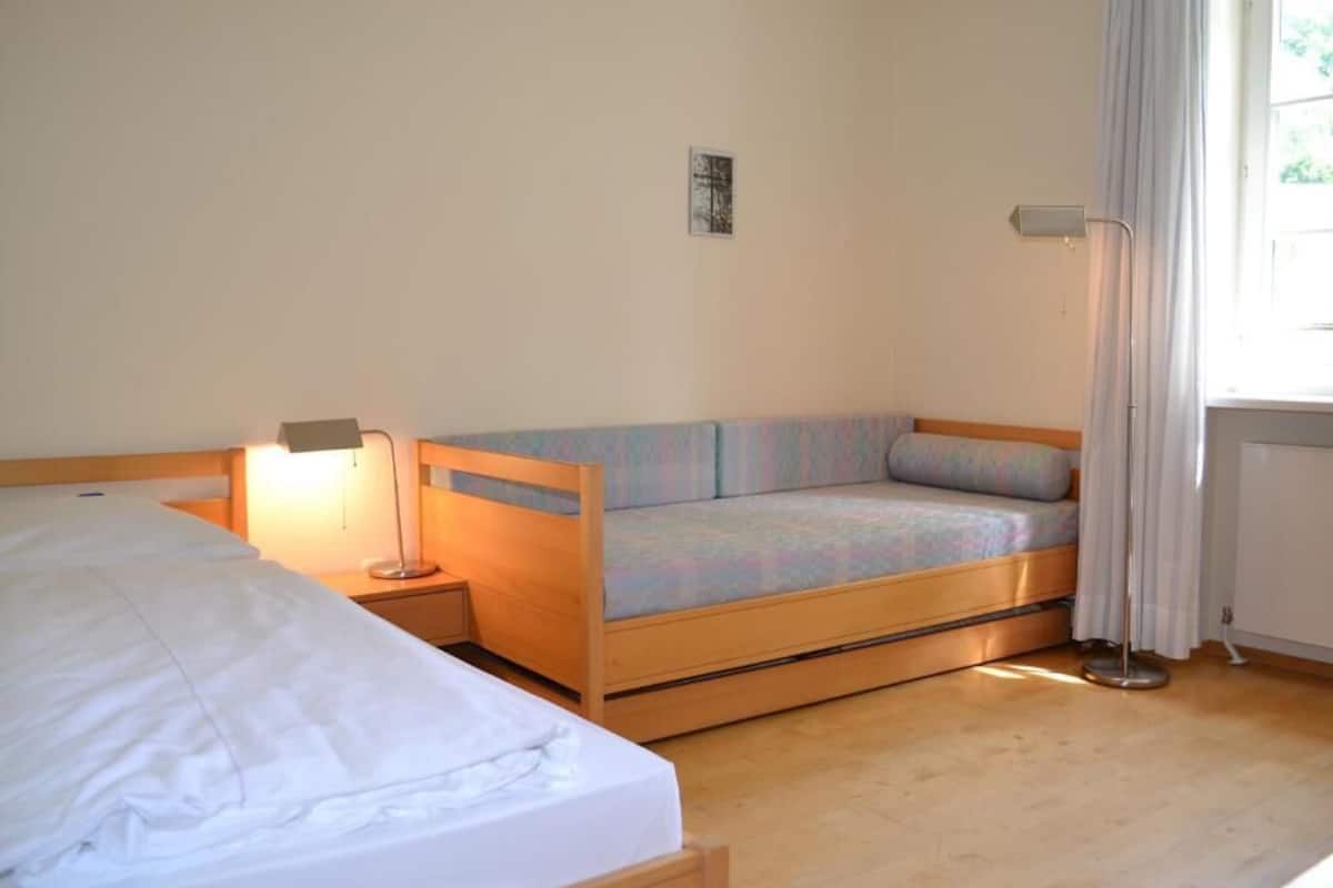 A small, cozy room with a bed, a sofa bed, a bedside table with a lamp, and a window with curtains.