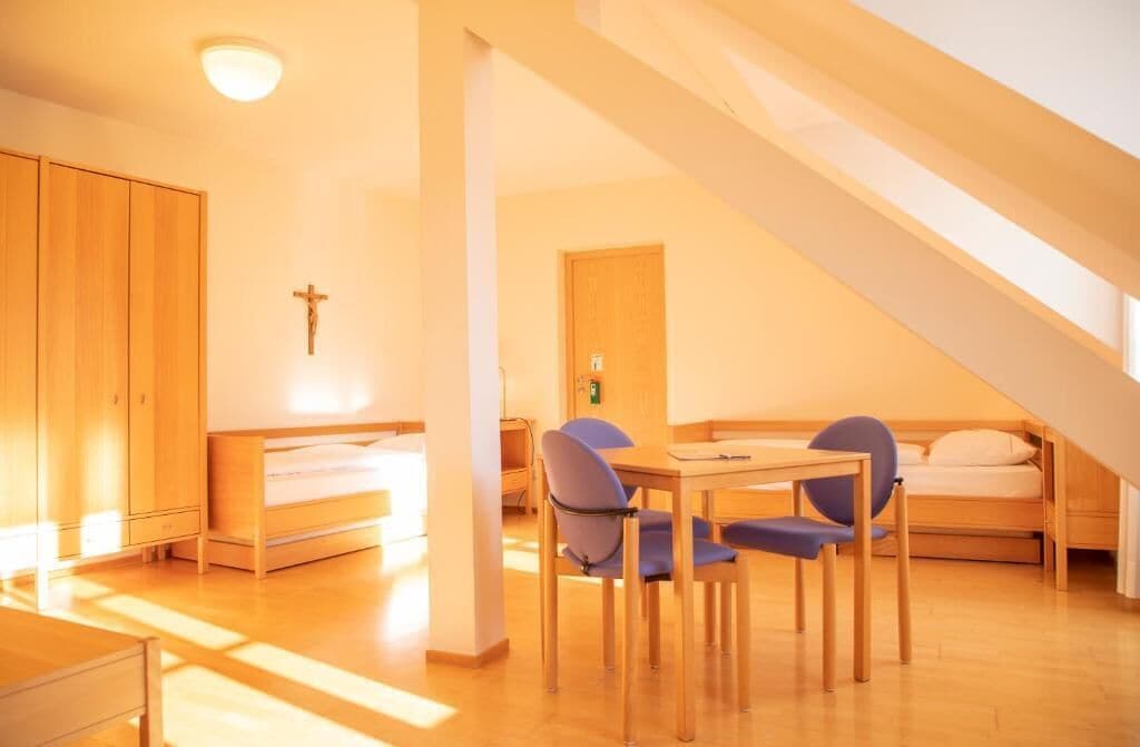 A room with a bed, a desk, and a chair, a cross on the wall, and a staircase.