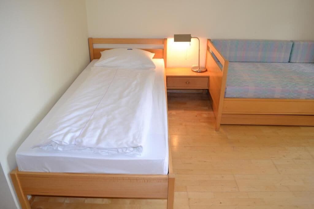 En säng för en person med en träram, ett sängbord med en lampa och en liten låda.