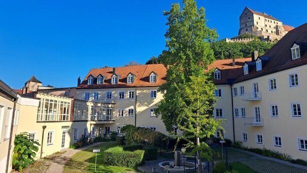 A courtyard with buildings, a tree, and a castle in the background. - Haus der Begegnung HEILIG GEIST (Burghausen)