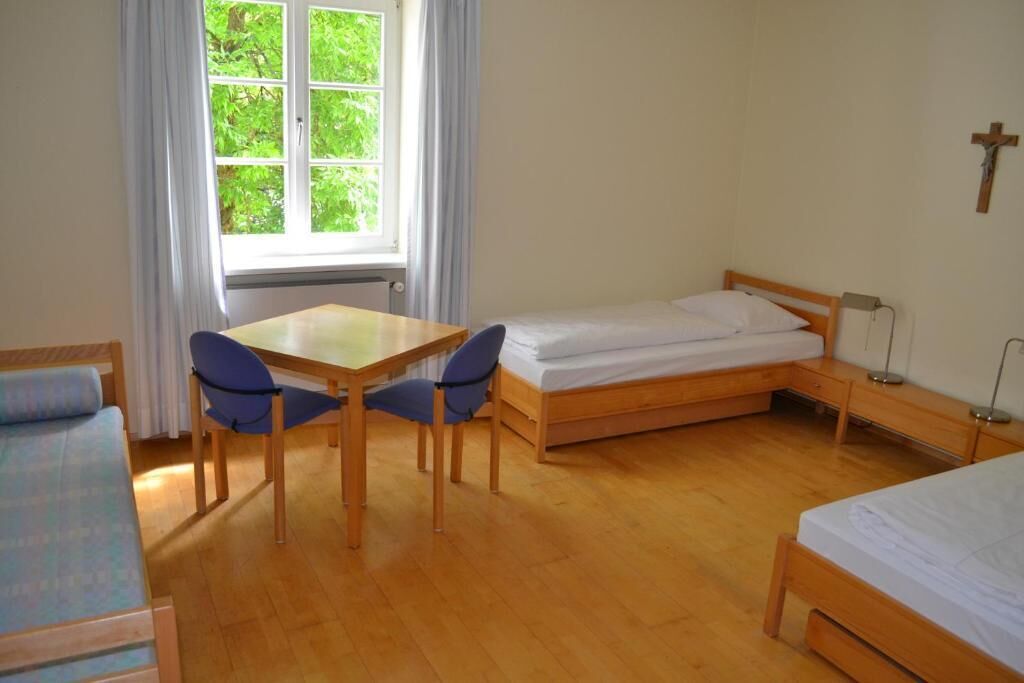 Una habitación con una cama individual, una mesita con dos sillas, un ventanal con cortinas y una cruz en la pared.