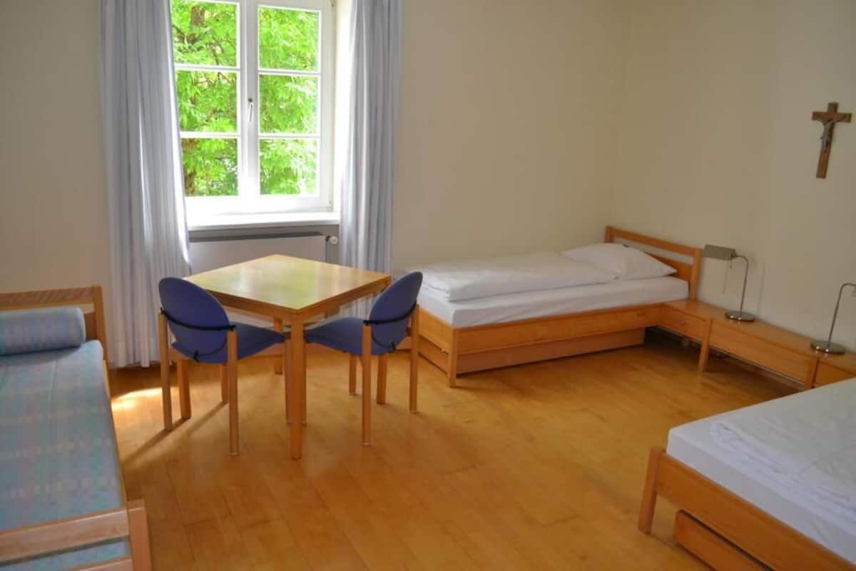 A room with a single bed, a small table with two chairs, a window with curtains, and a cross on the wall.