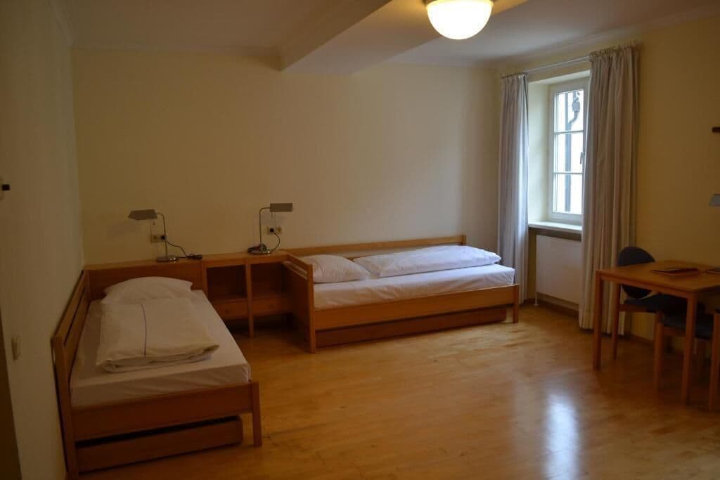 A room with two wooden beds, a small table, a chair, a lamp, and a window with curtains.
