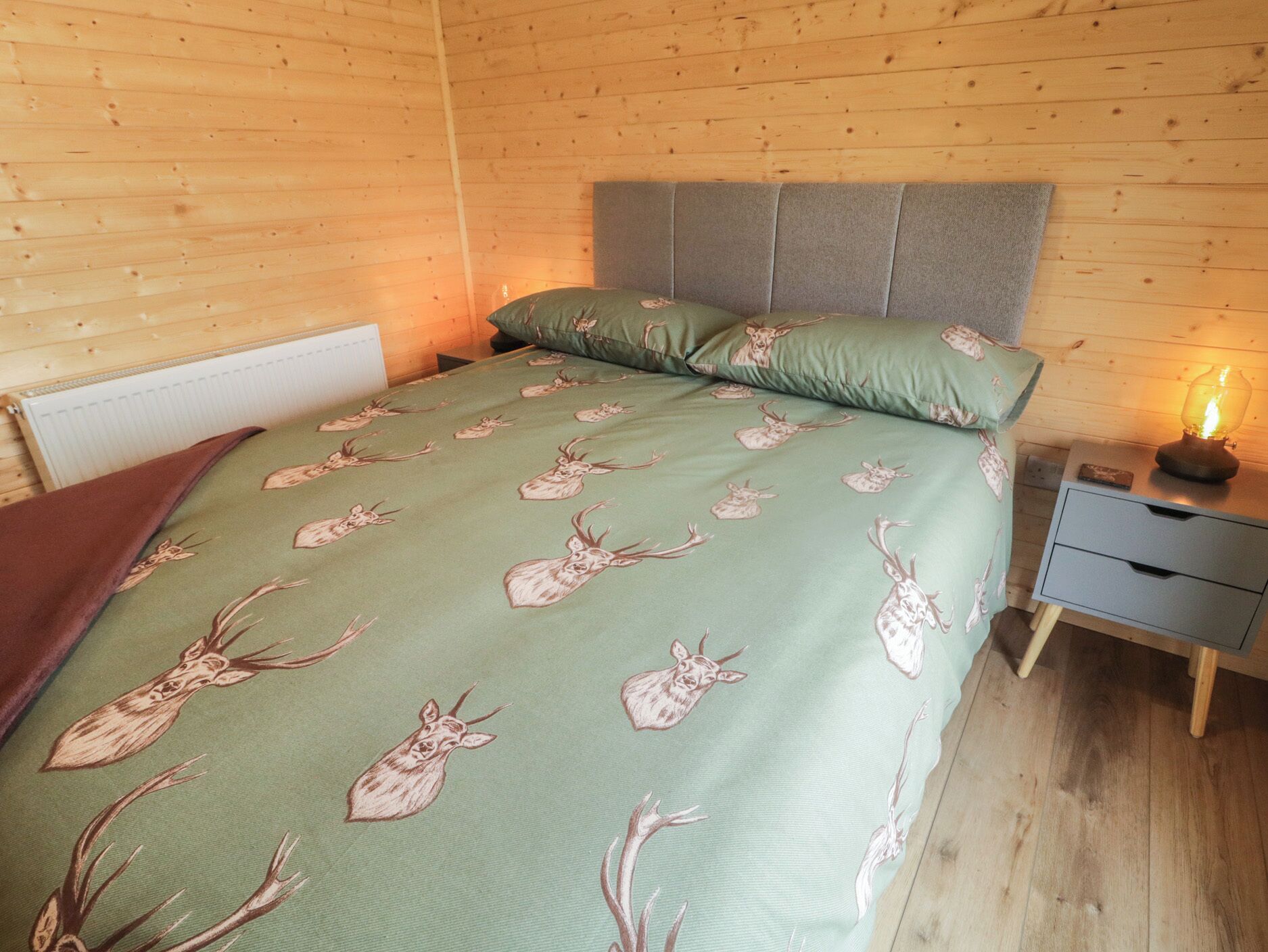 A wooden-paneled room with a bed featuring a green blanket with deer patterns, a grey headboard, and a bedside table with a lamp.