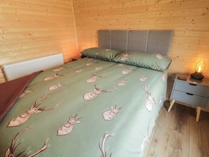 A wooden-paneled room with a bed featuring a green blanket with deer patterns, a grey headboard, and a bedside table with a lamp. - Alken The Stag (Bushmills)