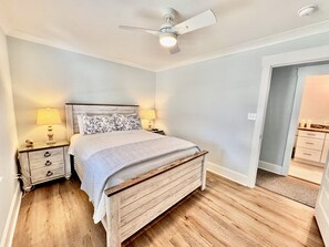 A bedroom with a wooden bed, two bedside tables with lamps, a ceiling fan, and a door leading to a bathroom. - Family Fun Getaway 4BR 2BA Short Walk To The Pier Outdoor Entertainment Space (Oak Island)