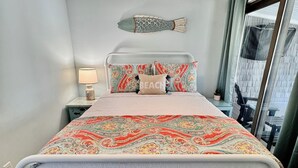 Ein ordentlich bezogenes Bett mit einem gemusterten Bettüberwurf, einer Nachttischlampe und einem Wandbild einer Fisch.'