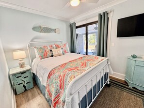 A bedroom with a bed, a nightstand, a lamp, a fish-themed rug, and a view of the outdoors. - Family Fun Getaway 4BR 2BA Short Walk To The Pier Outdoor Entertainment Space (Oak Island)