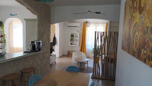 Zona pranzo con tavolo in legno, sedie e macchina da caffè. C'è un angolo cottura con parete in pietra e un cesto di frutta appeso.