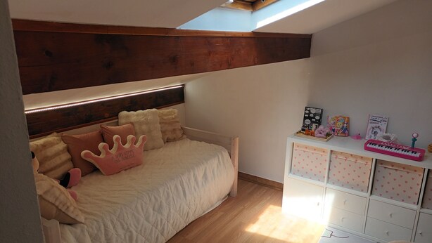 Una camera da letto con soffitto spiovente, un letto con testiera in legno, un lucernario e un comò bianco con cassetti.