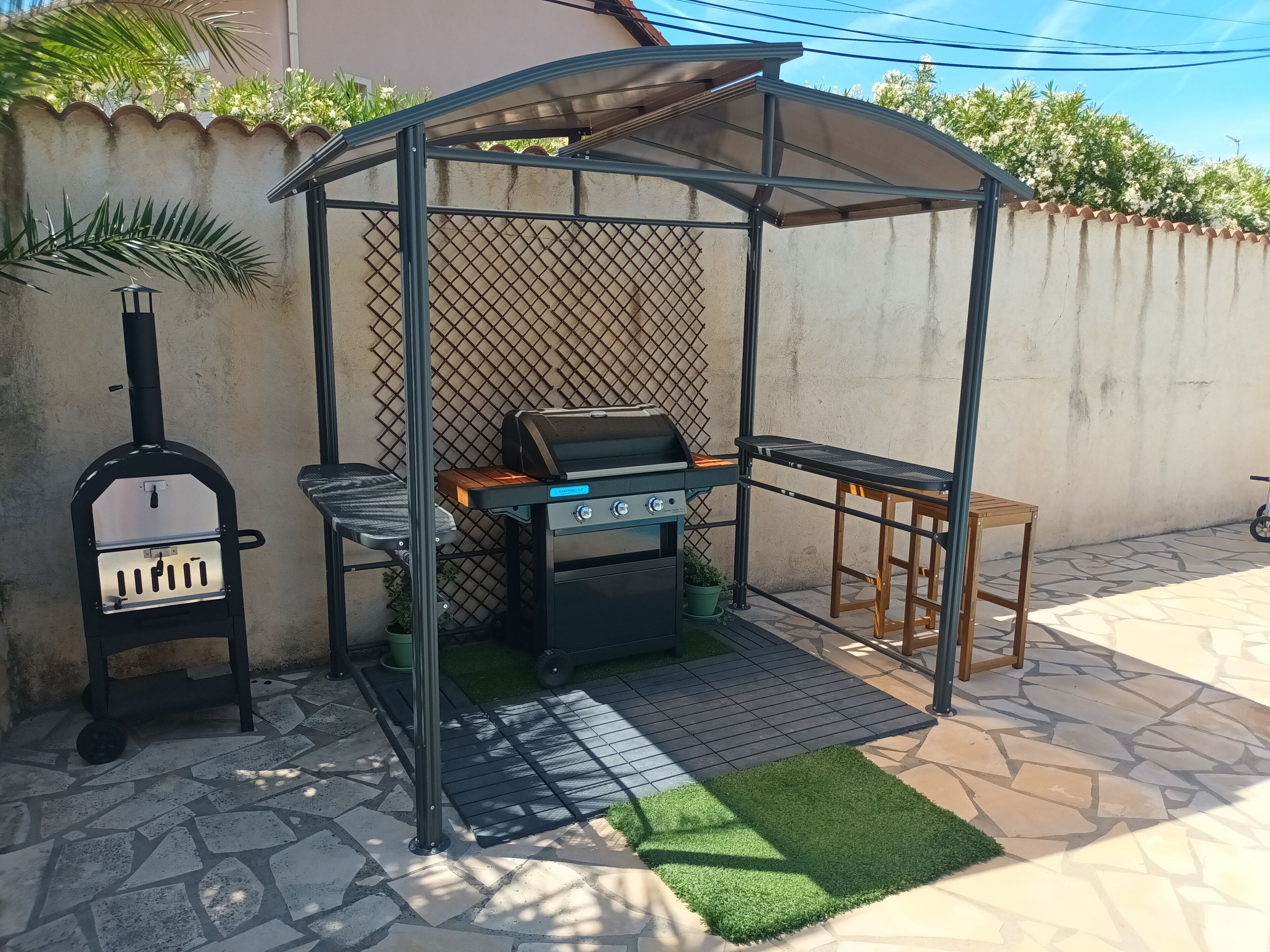 En uteplats med en grill, en pizzaugn och ett bord under en pergola.