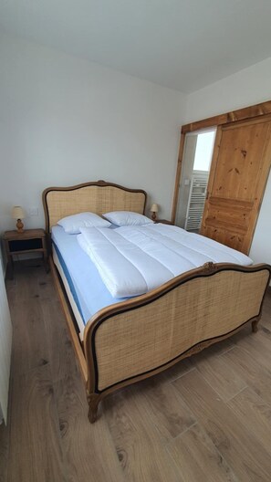 Una cama individual con estructura de madera y cabecero tejido, una mesita de noche con lámpara y un armario de madera.