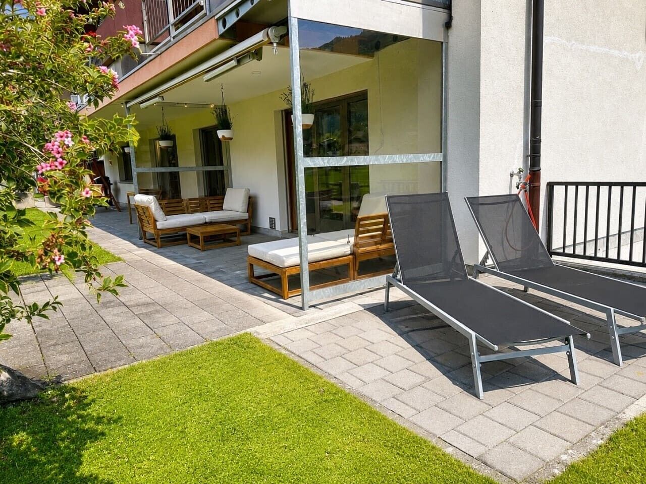 Un espace de détente extérieur moderne, doté d’une structure en verre, de mobilier en bois et d’une pelouse bien entretenue.