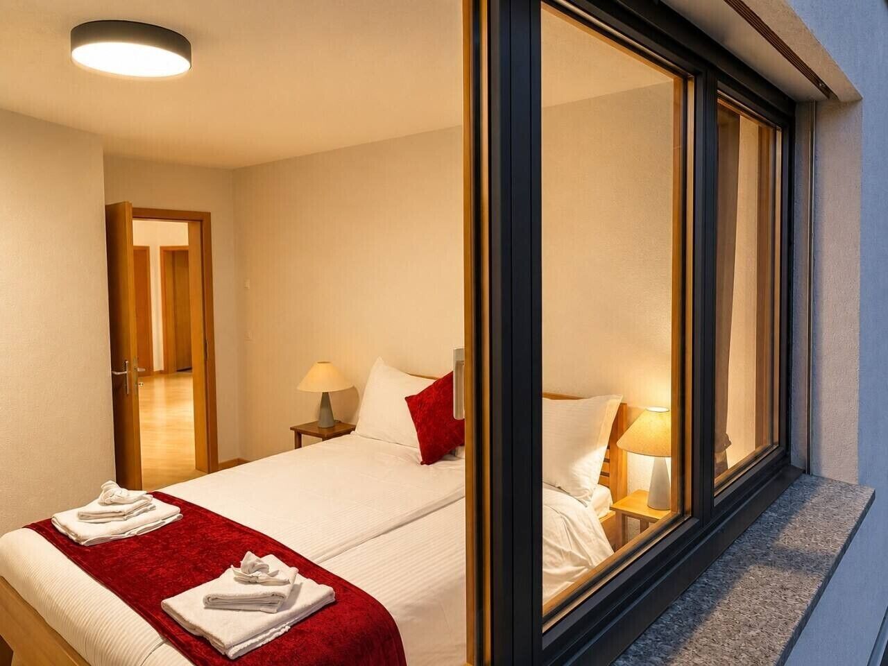A hotel room with a bed, bedside lamps, a window with a view, and a door leading to another room.
