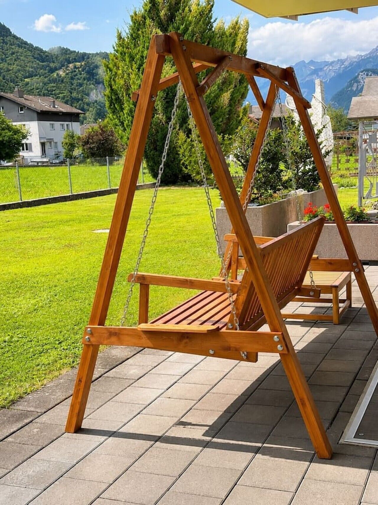 Um conjunto de baloiço de madeira com um banco, situado num alpendre com vista para um relvado verde e montanhas ao fundo.