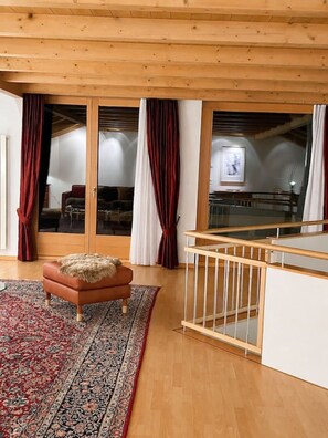 Een kamer met een houten plafond, een vloerkleed met een rood-wit patroon, een bruine leren bank en een houten leuning.