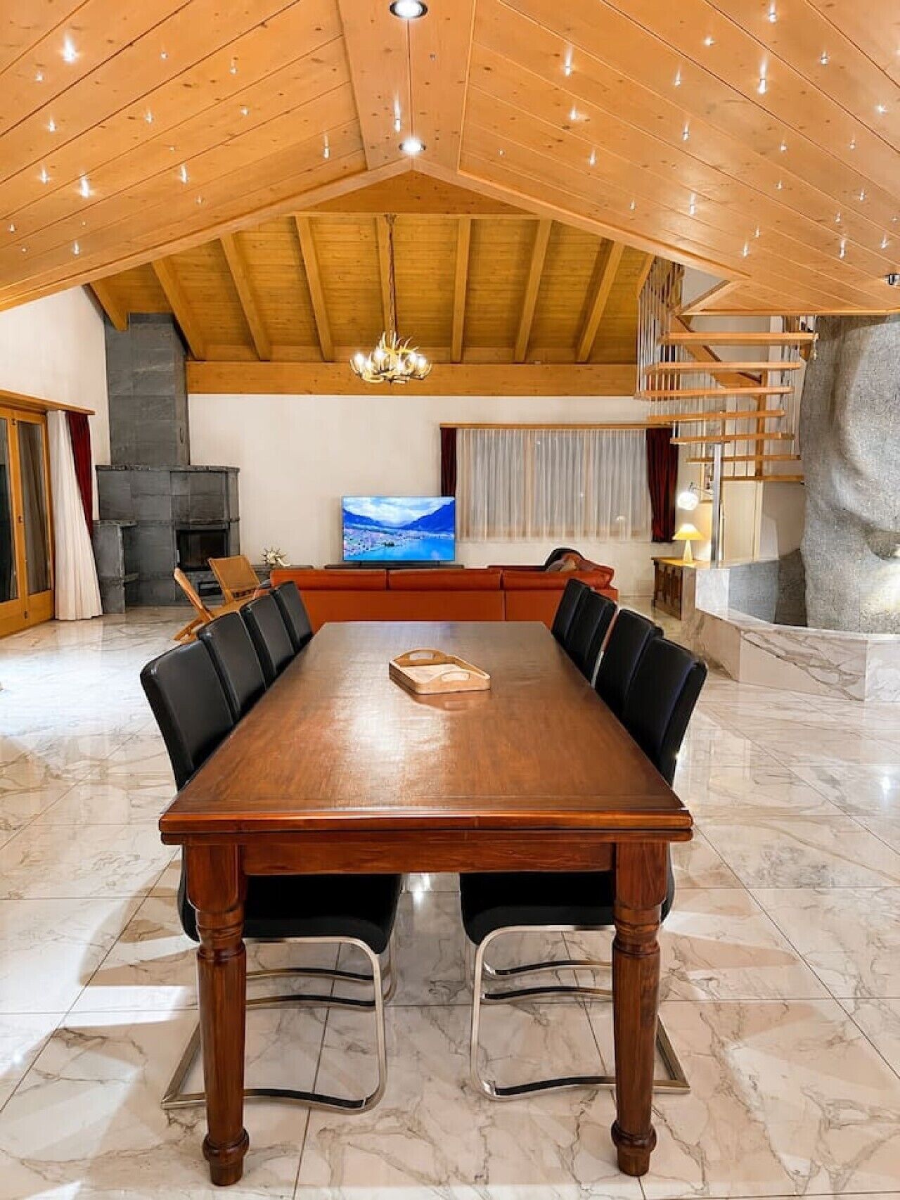 A spacious room with a large wooden dining table surrounded by chairs, a fireplace, a television, and a staircase.