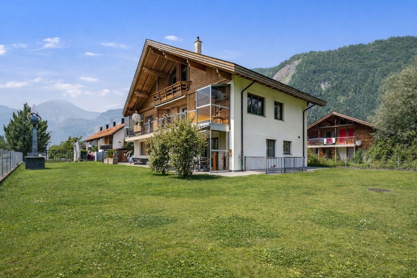 Un bâtiment à deux étages doté d’un balcon, d’une cour verdoyante et ayant une chaîne de montagnes en toile de fond.