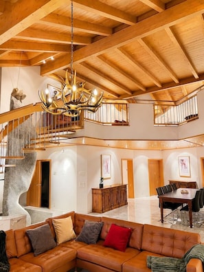 A spacious interior with a high ceiling, a grand chandelier, a staircase, a dining area, and a seating area.