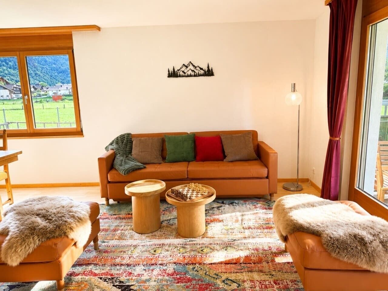 A living room with a sofa, two wooden side tables, a chessboard, a patterned rug, and a view of a green landscape through a window.