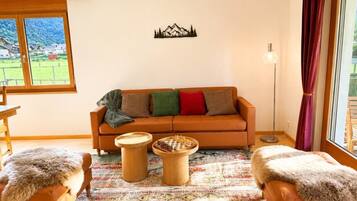 A living room with a sofa, two wooden side tables, a chessboard, a patterned rug, and a view of a green landscape through a window.