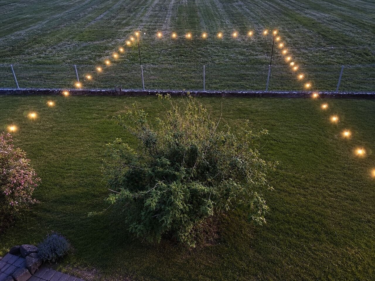 Un champ verdoyant, avec une haie au premier plan et des guirlandes lumineuses formant un cœur au loin.