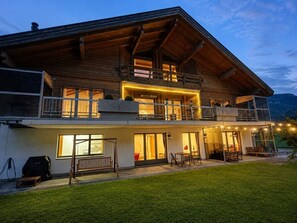 Een houten huis met twee verdiepingen, een balkon, grote ramen en een goed verlicht terras.