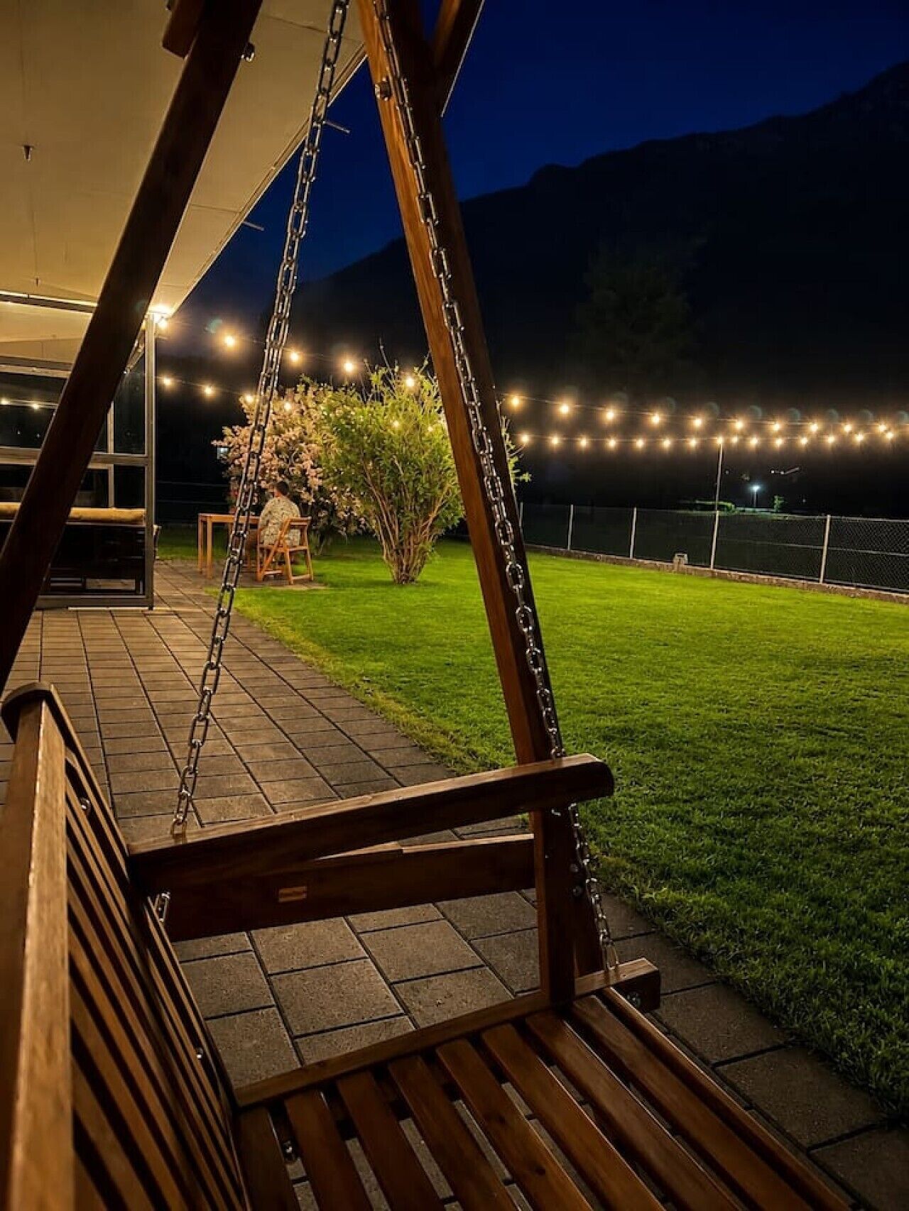 A wooden swing chair on a patio with a view of a grassy area and a fence.