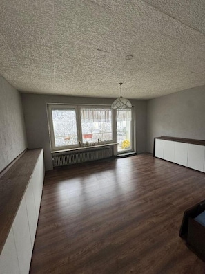 Et værelse med skråvæggede lofter, store vinduer, en radiator og en hængende lysarmatur.