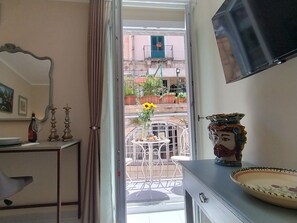 Una habitación con una puerta corredera que da a un balcón exterior, un televisor montado en la pared, una mesa con un jarrón de flores y una pieza decorativa de cerámica.