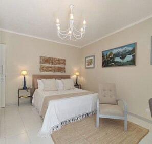 A bedroom with a bed, two bedside tables, a chair, a chandelier, and wall art. - Vrbo Property (Modica)