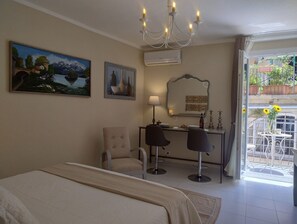 Hotelkamer met een bed, bureau, twee stoelen, een kroonluchter, schilderij, spiegel en balkon met potplanten.