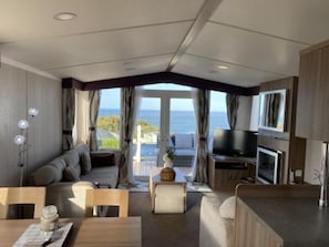 A modern RV interior with a sofa, dining table, TV, and a view of the ocean. - Cabin Sleeps 6 With Parking - Pets Welcome (Filey)